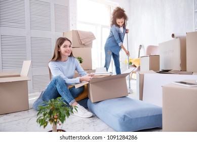 In Harmony. Pleasant Female Student Taking The Books Out Of The Box, Unpacking The Belongings, While Her Roommate Cleaning The Floor In Their New Dorm Room