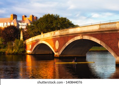 Harvard University bridge over the Charles River in early morning golden light, with a lone sculler passing under the span