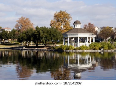 Harveston Lake Park in Temecula California. A white gazebo that is near a large body of water. Trees are placed in surrounding areas. Cloudy blue sky, and ripples on the lake.