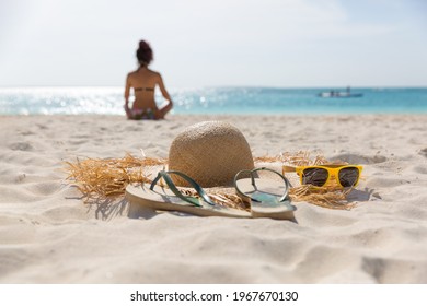 A hat with large brim, flip-flops and yellow sunglasses lie on the beach on the sand, against the background of the sea and the silhouette of a girl sitting on the sand, the girl is out of focus - Powered by Shutterstock