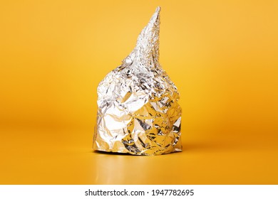 Hat Made Of Aluminium Foil On Yellow Background. Used By Those Who Are Afraid Of 5G Radiation Or Aliens. Symbol For Conspiracy Theory. Tin Foil Cap Paranoia