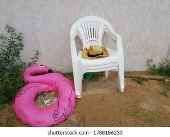 The Hat Is On A Plastic Chair Against A White Wall, And The Chair Is On The Sand. The Inflatable Flamingo Deflated Against The Wall.