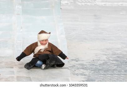 Hatgal, Mongolia, 4th March 2018: Mongolian Kid Dressed In Traditional Clothing On A Frozen Lake Khuvsgul, Coming Down A Slide