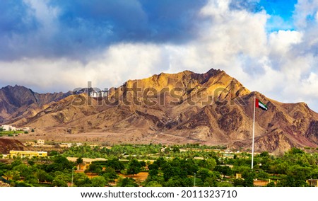 Hatta city welcoming sign written with large letters placed in Hajar mountains and UAE flag flying high in Hatta enclave of Dubai in the United Arab Emirates.