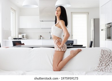 Haughty Proud Barefoot Young Woman In Summer Shorts Posing Straddling The Back Of A Sofa In A High Key Living Room With Hands On Hips Looking Down Thoughtfully At The Camera