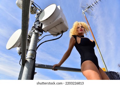 Haute Couture Portrait Of A Sophisticated Blonde Woman With Curly Hair On The City Rooftop In The Open Air, Among Communication And Telephone Antennas And Satellite Dishes. Urban Fashion Concept.