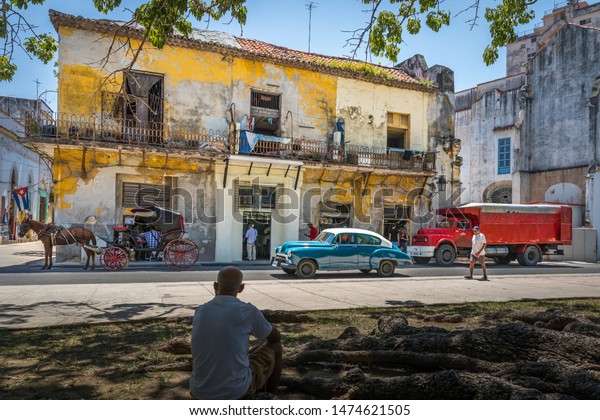 HAVANA 22 July 2018, typical colorful\
cityscape in Havana-Cuba with classic blue car, red truck, a horse\
and wagon and an old dilapidated yellow building with Cuban flag a\
man sitting in\
foreground