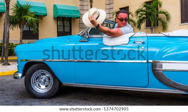 Havana Cuba, cab driver\
sitting in his old american car, hat down, waiving at some woman in\
front of a luxury hotel. Citizens of Havana are called Habanero in\
Spanish.