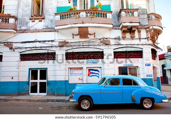 Havana, Cuba\
- December 11, 2016: Vintage classic american car parked in a\
street in front of a typical colonial house with a Fidel Castro\'s\
quote and cuba flag painting. Havana,\
Cuba.
