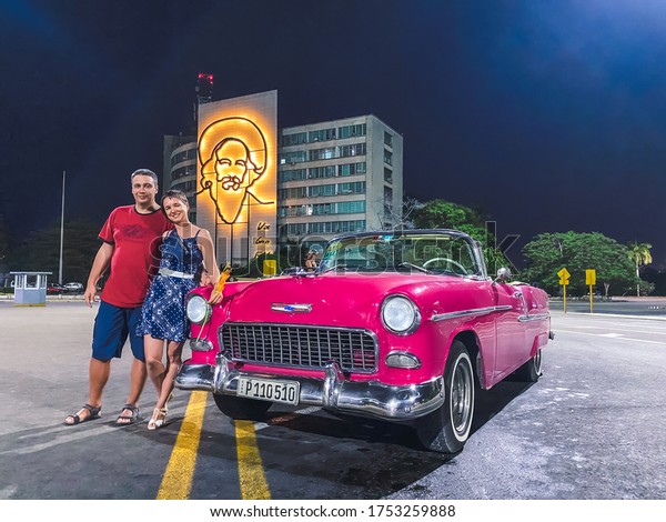 Havana, Cuba, February 2020: a young couple\
next to a pink retro car on Revolution Square, in the background a\
portrait of Camilo\
Cienfuegos