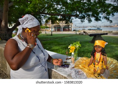 Havana, Cuba - June 04, 2015: A Doll Used In Santeria Practice By A Woman Talking On The Phone, In Regla Neighbourhood, Next To Some Flowers And A Bottle Of Water, A Fan, A Candle, In The Middle Of Th