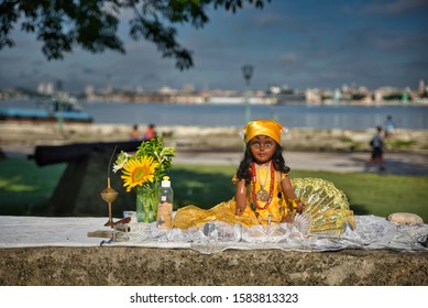 Havana, Cuba - June 04, 2015: A Doll Used In Santeria Practice By Women, In Regla Neighbourhood, Next To Some Flowers And A Bottle Of Water, A Fan, A Candle, In The Middle Of The Day, In Havana, Cuba,