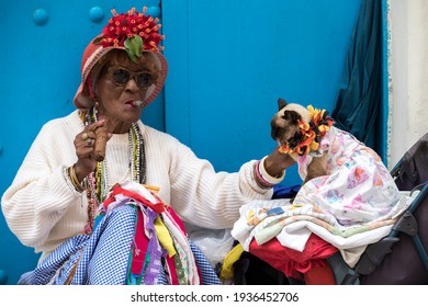 HAVANA, CUBA - MARCH 2, 2021: An Older Cuban Woman Smokes Two Cigars And Pets A Cat