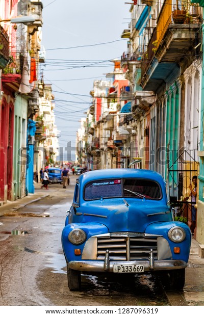 HAVANA, CUBA - NOVEMBER 14, 2017: Typical\
street scene with people, old cars and colorful buildings. With\
over 2 million inhabitants Havana is the capital of Cuba and the\
largest city in the\
Caribbean