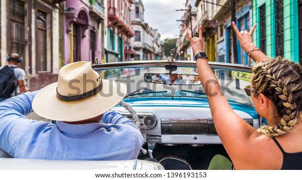 Havana Cuba. Woman smiling happy\
enjoying car road trip travel vacation, holding hands up in blue\
vintage convertible car on the colourful streets of\
Havana.