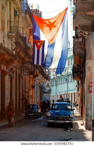 HAVANA,CUBA -\
SEPTEMBER 29,2018 : Urban scene with cuban flags, antique cars,\
people and aged buildings in Old\
Havana