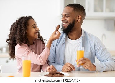 Have A Bite. Portrait of African American little girl feeding her happy daddy with homemade cookie. Smiling black man and his daughter having breakfast at home in the kitchen, drinking orange juice