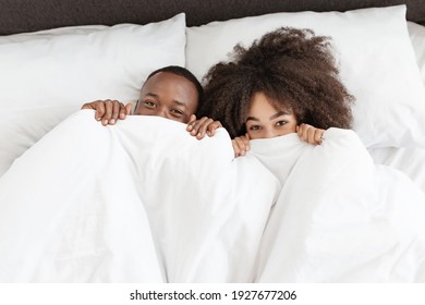 Have Fun In Bed Together During Free Time, Vacation Or Weekend. Funny Cute Millennial African American Husband And Wife Peeking Out From Under White Cover, In Cozy Bedroom, Top View, Copy Space