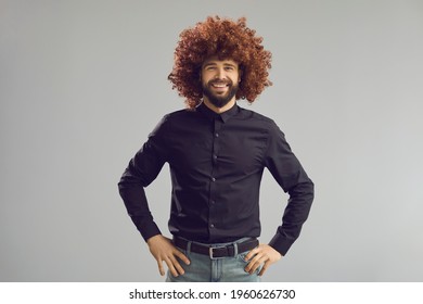 Have A Good Hair Day. Portrait Of A Positive Guy With A Crazy Hairstyle. Funny Happy Young Man Wearing A Curly Brown Wig Standing Hands Oh Hips On A Gray Studio Background