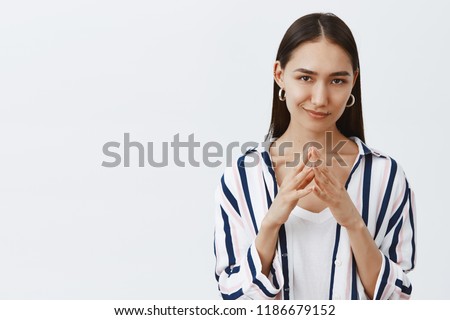I have great evil plan. Good-looking confident and tricky woman in striped blouse, holding fingers together and smirking at camera, having some bad intention in mind, planning to commit crime