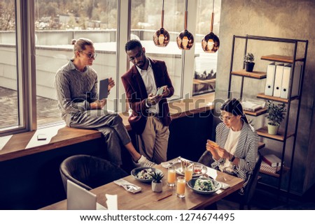 Have a look. Serious dark-skinned man standing near window and communicating with his partner