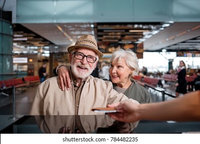 Have Nice Trip. Portrait Of Cheerful Gray-haired Couple Is Standing At International Airport Near Registration Desk. Old Man Is Taking His Tickets While Aged Woman Is Looking At Him With Love