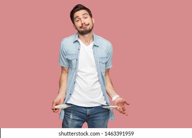 I have no money more. Portrait of sad bankrupt bearded young man in blue casual style shirt standing and showing his empty pocket and looking at camera. indoor studio shot, isolated on pink background