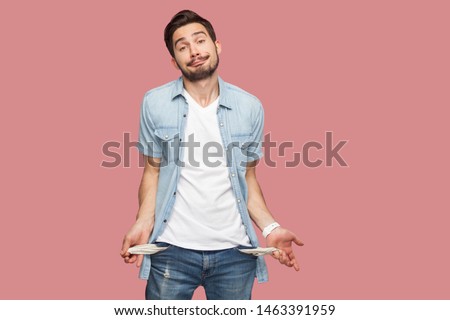 I have no money more. Portrait of sad bankrupt bearded young man in blue casual style shirt standing and showing his empty pocket and looking at camera. indoor studio shot, isolated on pink background