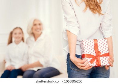 I Have Something For Both Of You. Selective Focus On A Turned Back Woman In Casual Looking At Her Daughter And Mom And Hiding A Beautifully Wrapped Present Behind Her Back.