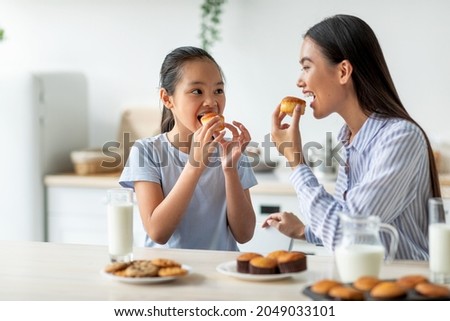 Having a bite. Cute asian girl and her mother eating snacks in kitchen, happy woman and her adorable female child enjoying homemade muffins and drinking milk together