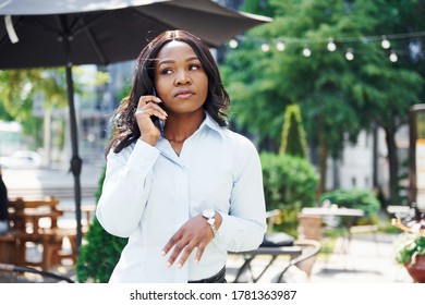 Having conversation by using phone. Young afro american woman in white shirt outdoors in the city near green trees and against business building.