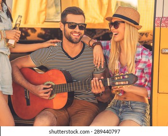 Having fun after long road trip. Handsome young man sitting in minivan and playing guitar while two young women bonding to him   - Powered by Shutterstock