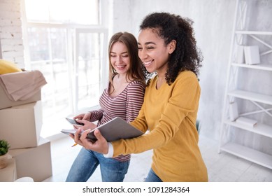 Having Fun. Beautiful Female Students Creating The List Of Boxes In The Notebook And Laughing Before Moving Out Of The Dorm Room