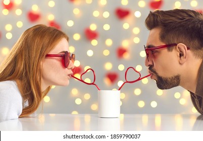 Having a fun date on Saint Valentine's Day. Young man and woman leaning over cafe table and sipping drink from one cup through heart-shaped straws enjoying cute and funny couple moment