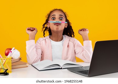 Having Fun. Funny black girl holding pencil between nose and lips as mustache, taking break from study and homework, sitting at desk with laptop isolated over yellow studio wall, banner