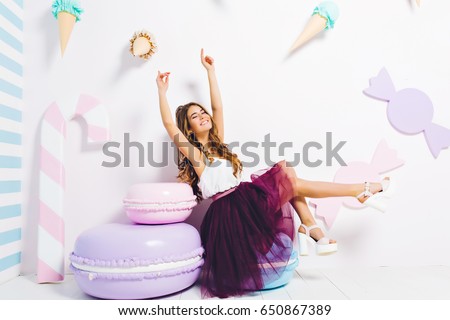 Having fun of joyful amazing model in purple tulle skirt on white heels expressing on big macaroon isolated on white background surround big sweets. Beautiful young woman in sweet dreams