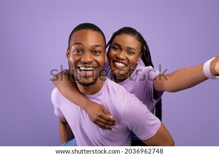 Having Fun. Portrait of happy black couple talking selfie, cheerful woman piggyback riding on her boyfriend's back, holding looking at camera standing isolated over purple studio wall