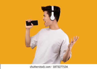 Having Fun. Portrait Of Overjoyed Asian Guy Singing Song And Using Smart Phone As Microphone, Wearing Wireless Headset. Carefree Young Man Dancing Isolated Over Orange Studio Background