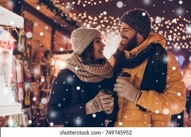 Having fun together at a christmas fairy with snowfall. Young cheerful couple is having a walk with hot drinks, enjoying, dressed warm, looking at each other and laugh, snowflakes all around - Shutterstock ID 749745952