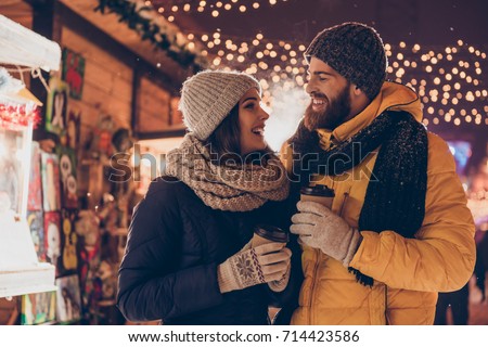 Having fun together at a christmas fairy. Young cheerful couple is having a walk with hot drinks, enjoying, dressed warm, looking at each other and laugh