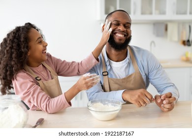 Having Fun While Cooking Concept. Portrait Of Happy African American Dad And His Child Daughter Baking In Kitchen And Fooling Around, Playful Girl Slapping Father's Face With Flour Palm