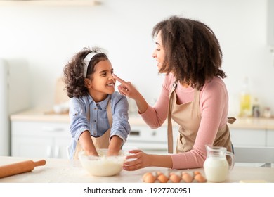 Having Fun While Cooking Concept. Portrait of happy african american mom and her child daughter baking in kitchen and fooling around, woman playfully touching girl's nose, kneading dough - Powered by Shutterstock