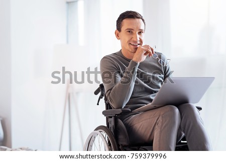 Having secret. Enigmatical male keeping smile on his face and holding glasses in right hand while sitting on the wheelchair