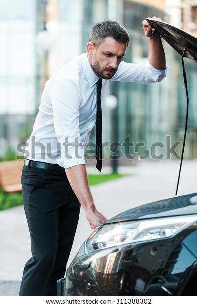 Having some car\
troubles. Serious young businessman looking inside of the vehicle\
hood while standing near his\
car