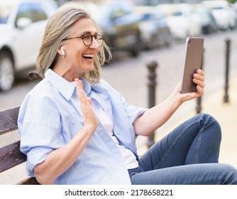 Having A Video Call Using Digital Tablet And Earphones Mature Woman With Grey Hair Sitting On The Bench At The Streets Of Old European Town. Mature Woman In Denim And Blue Shirt Outdoors.