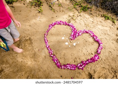 Hawaii love honeymoon. Heart shape lei flowers on sand beach for Hawaii honeymoon romantic vacation getaway travel. Pink orchids flower necklace lying on sand with legs of a person standing  by. - Powered by Shutterstock