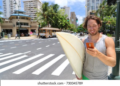 Hawaii Surfer Man Honolulu City Lifestyle Using Smart Phone Going To Waikiki Beach Surfing Holding Surfboard On Street. American Living In USA.