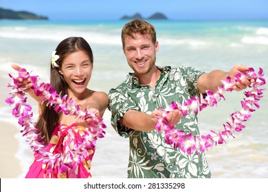 Hawaii Welcome - Hawaiian People Showing Leis Flower Necklaces As A Welcoming Gesture For Tourism. Travel Holidays Concept. Asian Woman And Caucasian Man On White Sand Beach In Aloha Clothing.