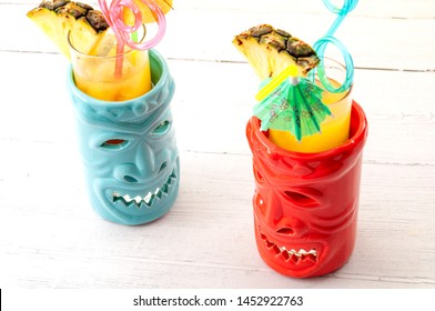 Hawaiian Bar, Summer Vacation And Beach Drinks Concept Theme With Tiki Mugs On White Wood Table With Each Mug Having A Plastic Straw, A Cocktail Umbrella And A Piece Of Pineapple