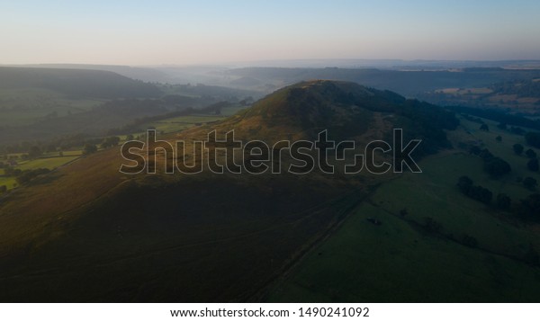 Hawnby Hill divides light and dark at sunrise in the\
beautiful British countryside, mist fading away in the background\
as the warm glow of the summer sun hits the valley under a hazy\
clear blue sky.